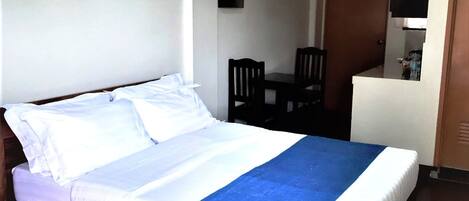 A hotel room with a wooden bed, a small table with two chairs, a white wall, and a door leading to a kitchenette.