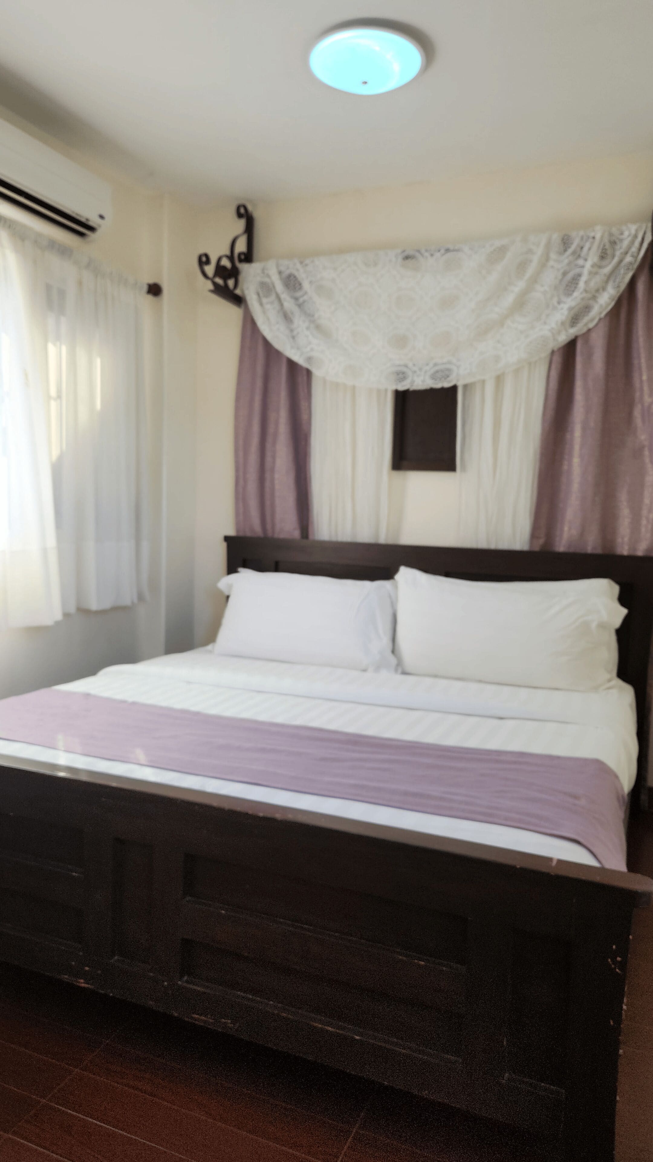 A bedroom with a wooden bed, white bedding, a window with white and purple curtains, and a ceiling light.