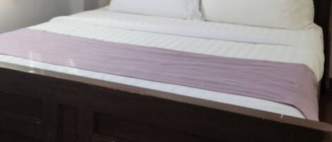 A bedroom with a wooden bed, white bedding, a window with white and purple curtains, and a ceiling light.