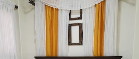 A bed with white linens, a yellow blanket, and a wooden headboard. There is a white lace curtain and a wooden frame with a picture above the bed.