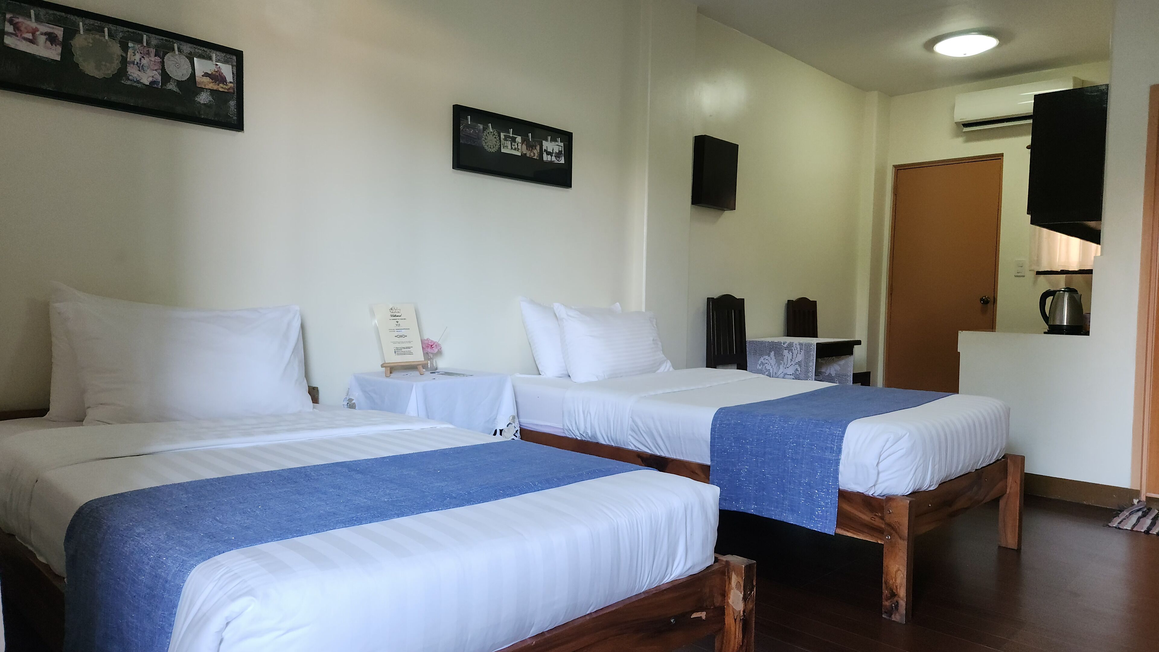 A hotel room with two single beds, a small table with a sign, a mirror, a wardrobe, and a kitchenette area.