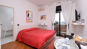 Une chambre avec une couvre-lit rouge, une petite table de chevet, une fenêtre avec des rideaux, une vue sur la ville et un balcon avec une table et des chaises.
