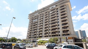 Un immeuble à plusieurs étages doté de nombreux balcons, un parking où sont garées diverses voitures et un ciel dégagé.