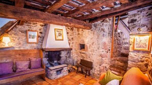 A rustic stone-walled room with a fireplace, wooden beams, a cushioned bench, and framed pictures on the walls.