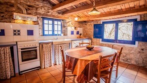 A rustic kitchen with stone walls, wooden beams, a round table with chairs, a stove, and a window with blue shutters.