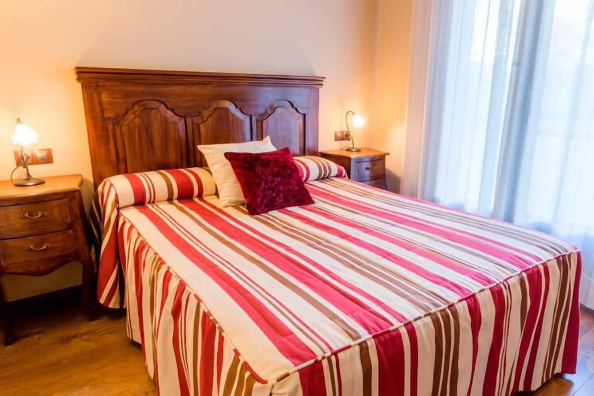 Een hotelkamer met een groot bed, een houten hoofdeinde, twee nachtkastjes met lampen, een bijzettafeltje en een raam met gordijnen.