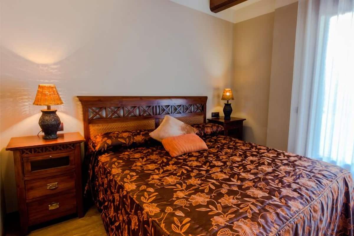 Een slaapkamer met een bloemenbedekking, een houten hoofdbord, twee nachtkastjes met lampen, een bijzettafeltje en een raam met gordijnen.