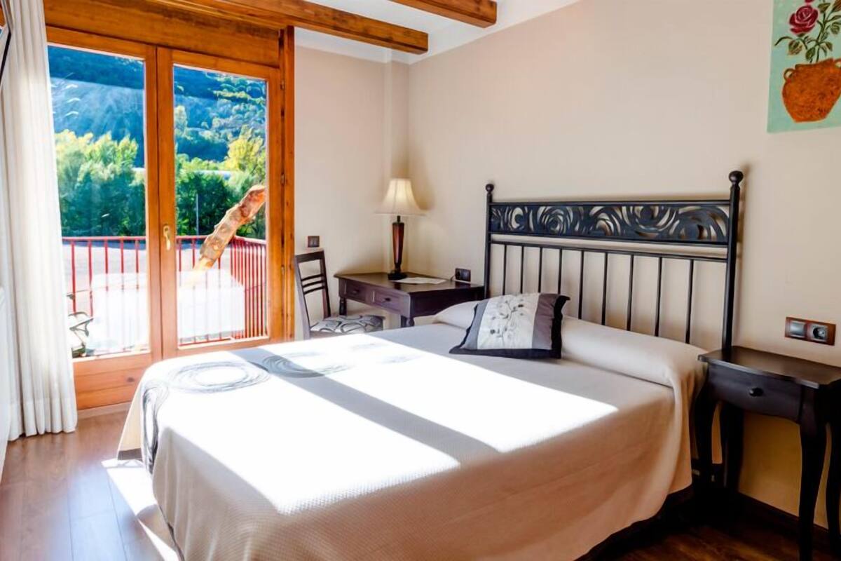 Een slaapkamer met een bed, een nachtkastje, een lamp en uitzicht op een balkon met een hekwerk.