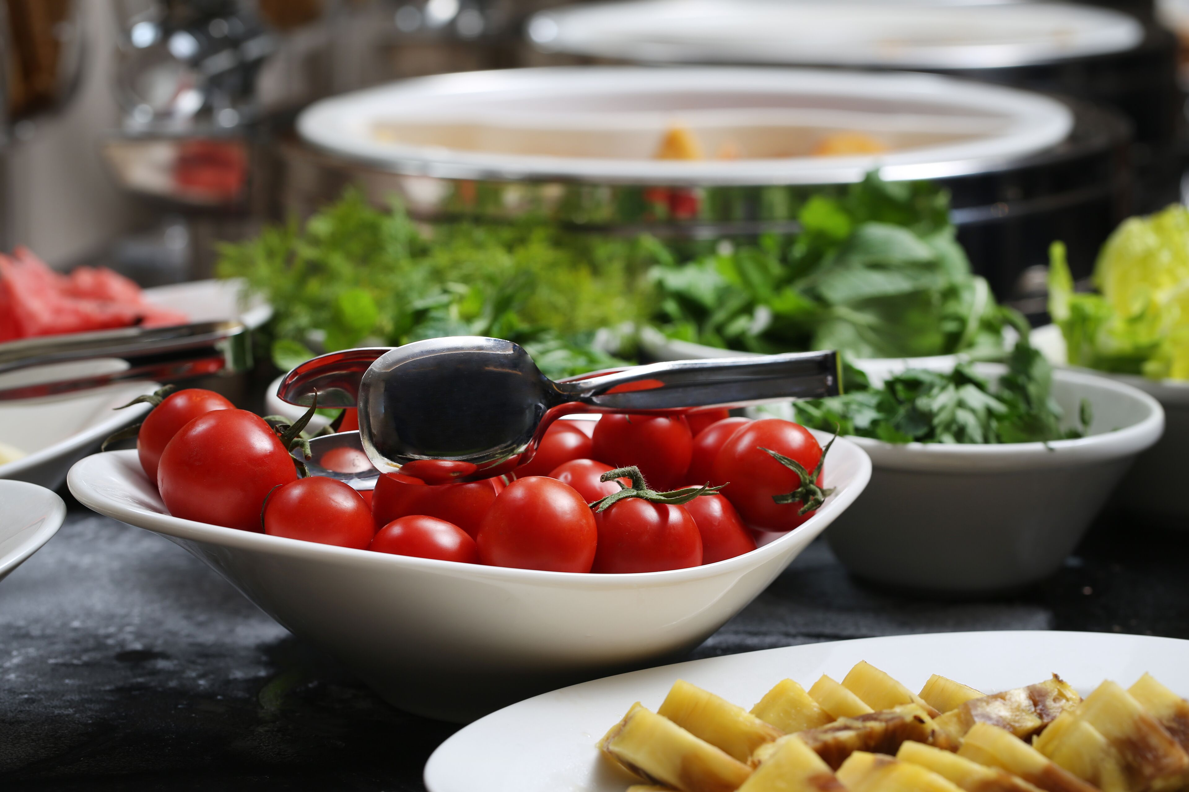 A buffet table with a bowl of tomatoes, a plate of sliced potatoes, and a bowl of greens.