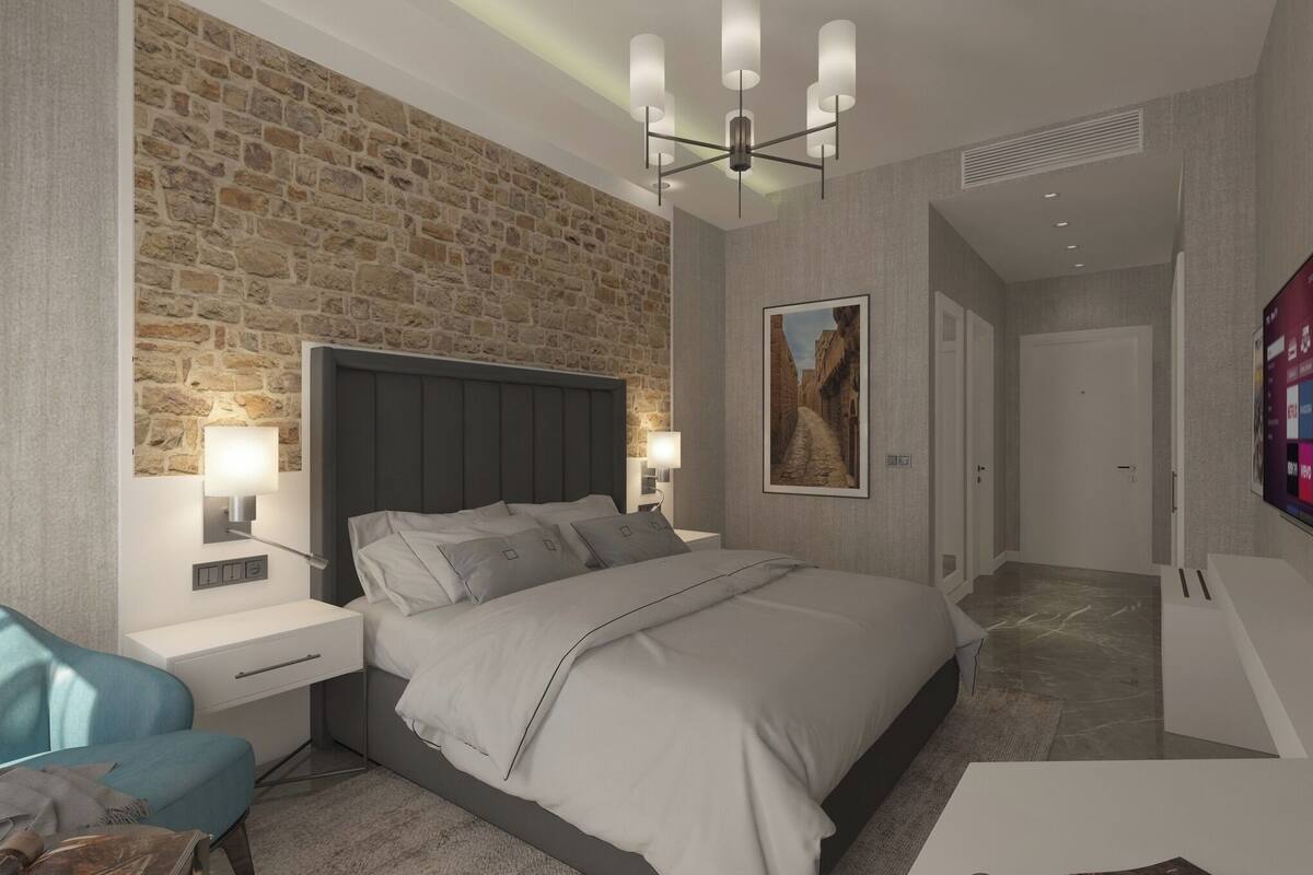 A modern bedroom with a stone wall, a large bed, bedside lamps, a TV, and a small seating area.