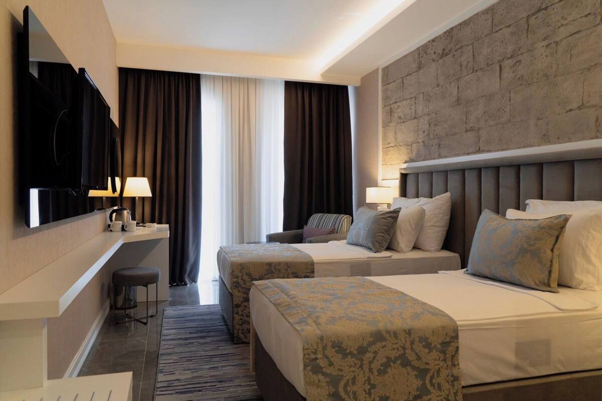 A hotel room with a large bed, a smaller bed, a desk with a chair, a television, and a view of the outside through a window with curtains.