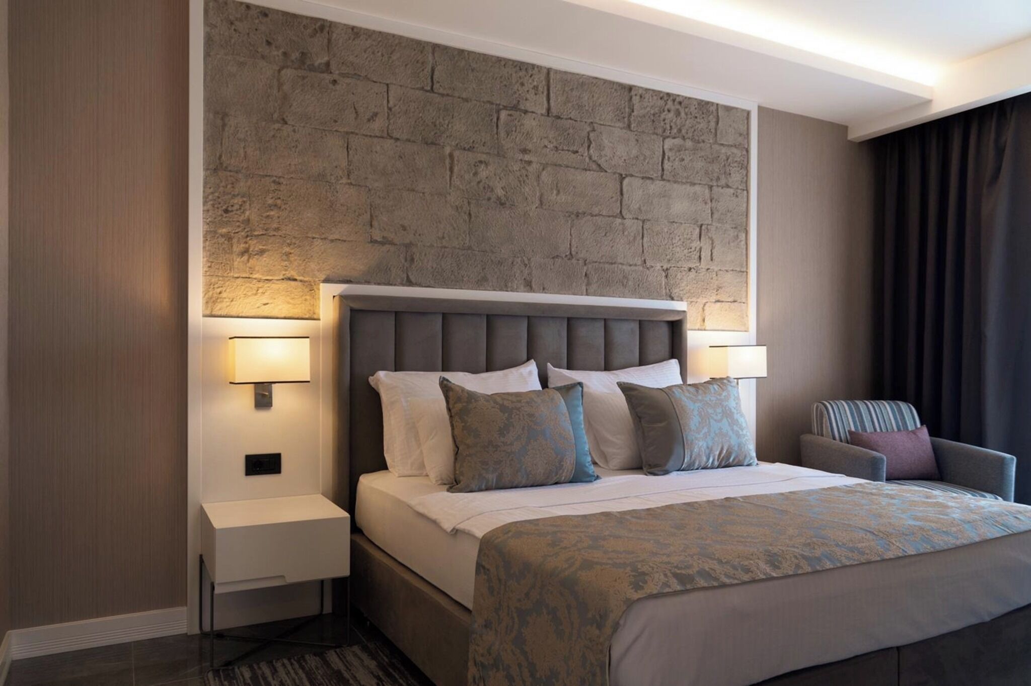 A hotel room with a bed, two bedside tables, a chair, a stone wall, and a window with curtains.