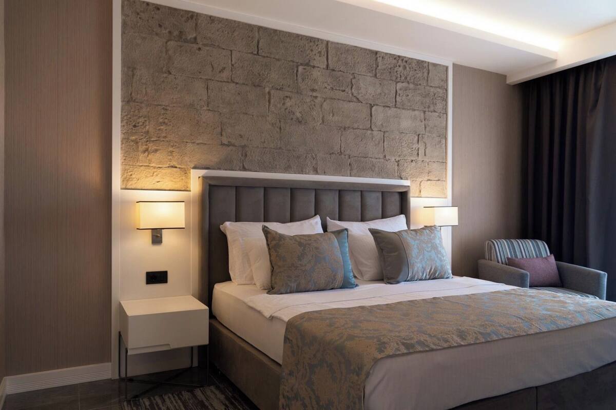 A hotel room with a bed, two bedside tables, a chair, a stone wall, and a window with curtains.