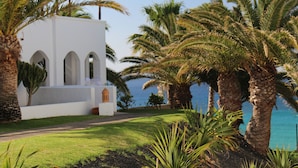 A white building with arched windows and a small tower, surrounded by palm trees and a clear view of the ocean. - Esencia de Fuerteventura by Princess - Adults Only (Pajara)