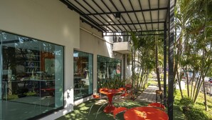An outdoor café area with glass walls, red tables, and chairs, surrounded by greenery and palm trees. - At Border (Aranyaprathet)