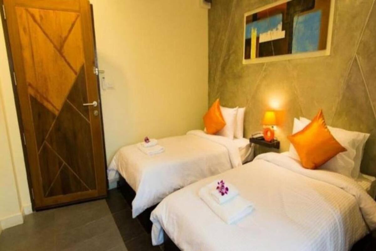 A hotel room with two beds, each with white bedding and orange pillows, a wooden door, and a nightstand with a lamp.