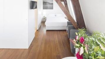 A modern loft bedroom with exposed wooden beams, a white bed, and a grey sofa.