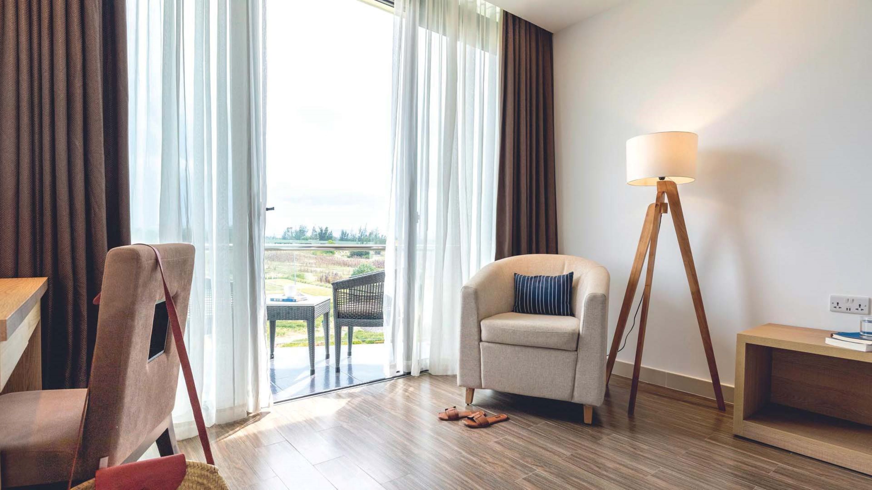 Una sala moderna con un sofá, una silla, una lámpara de pie y vistas a un jardín a través de amplios ventanales.