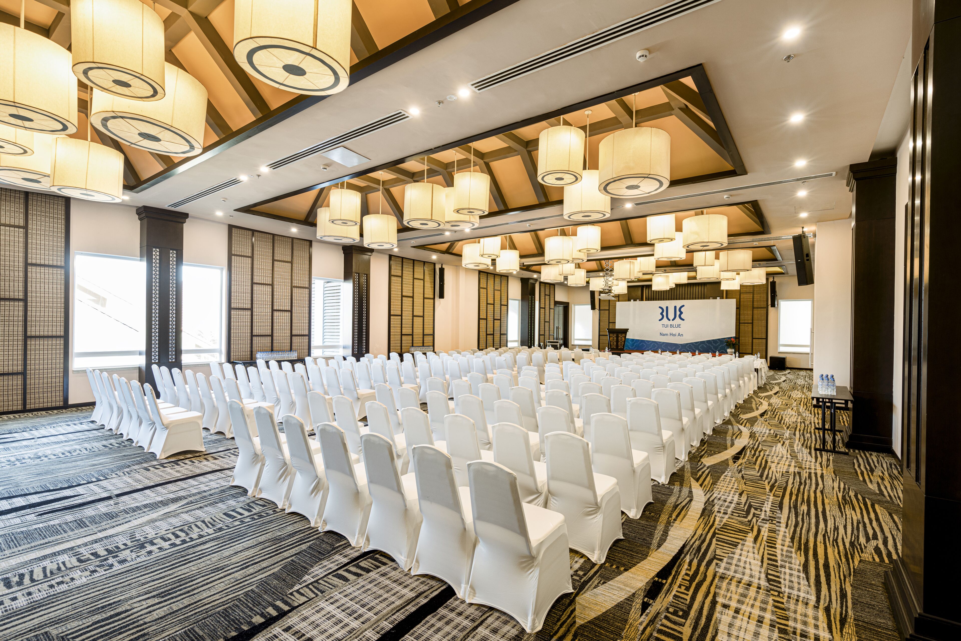 A conference room with rows of white chairs, a stage with a presentation screen, and a patterned carpet.