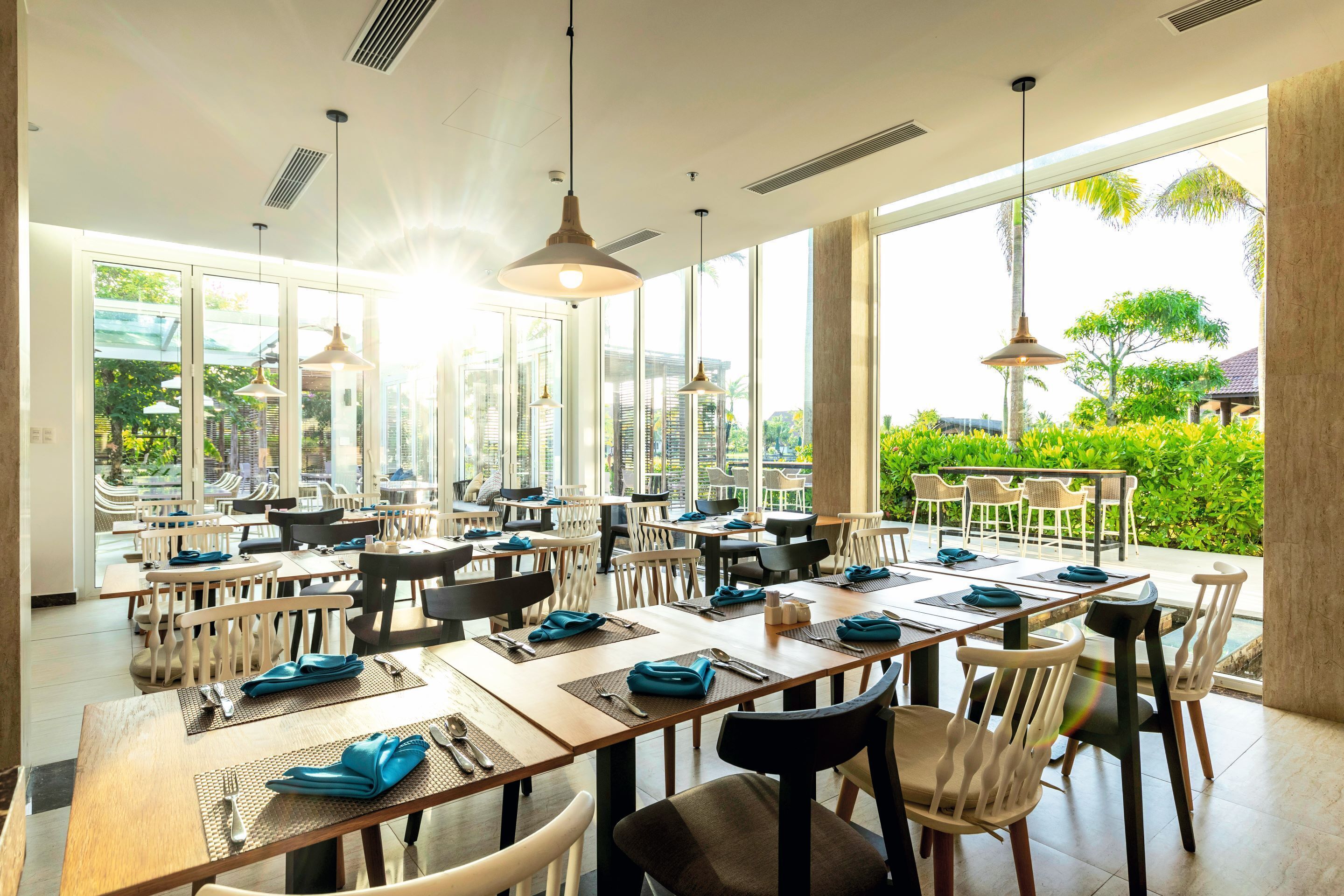 A spacious dining area with multiple tables and chairs, large windows offering views of greenery and buildings, and pendant lighting.