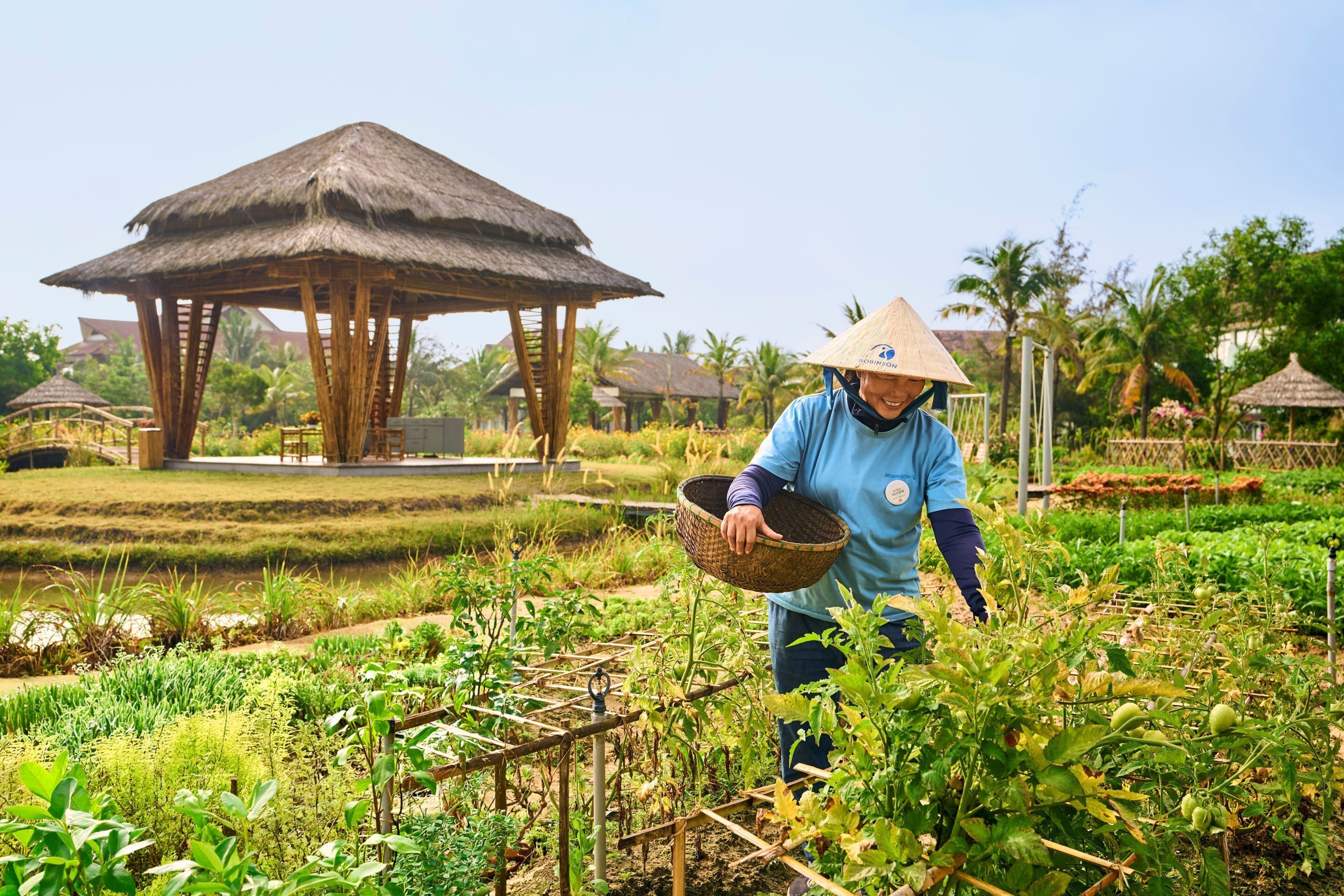 A person in a blue shirt and straw hat working in a garden with a thatched-roof structure in the background.