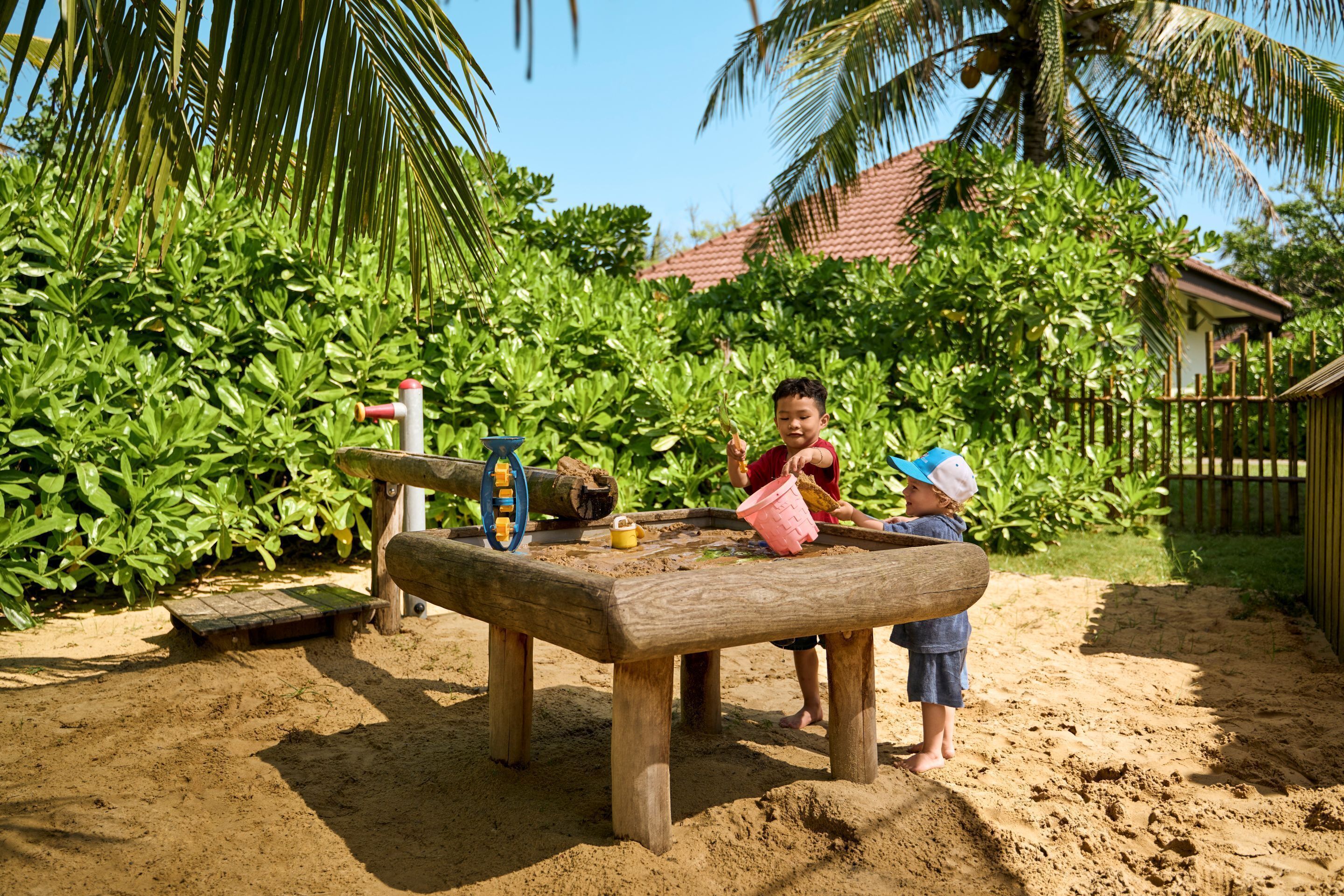 A wooden seesaw with a child on each side, set in a sandy area with greenery and a house in the background.
