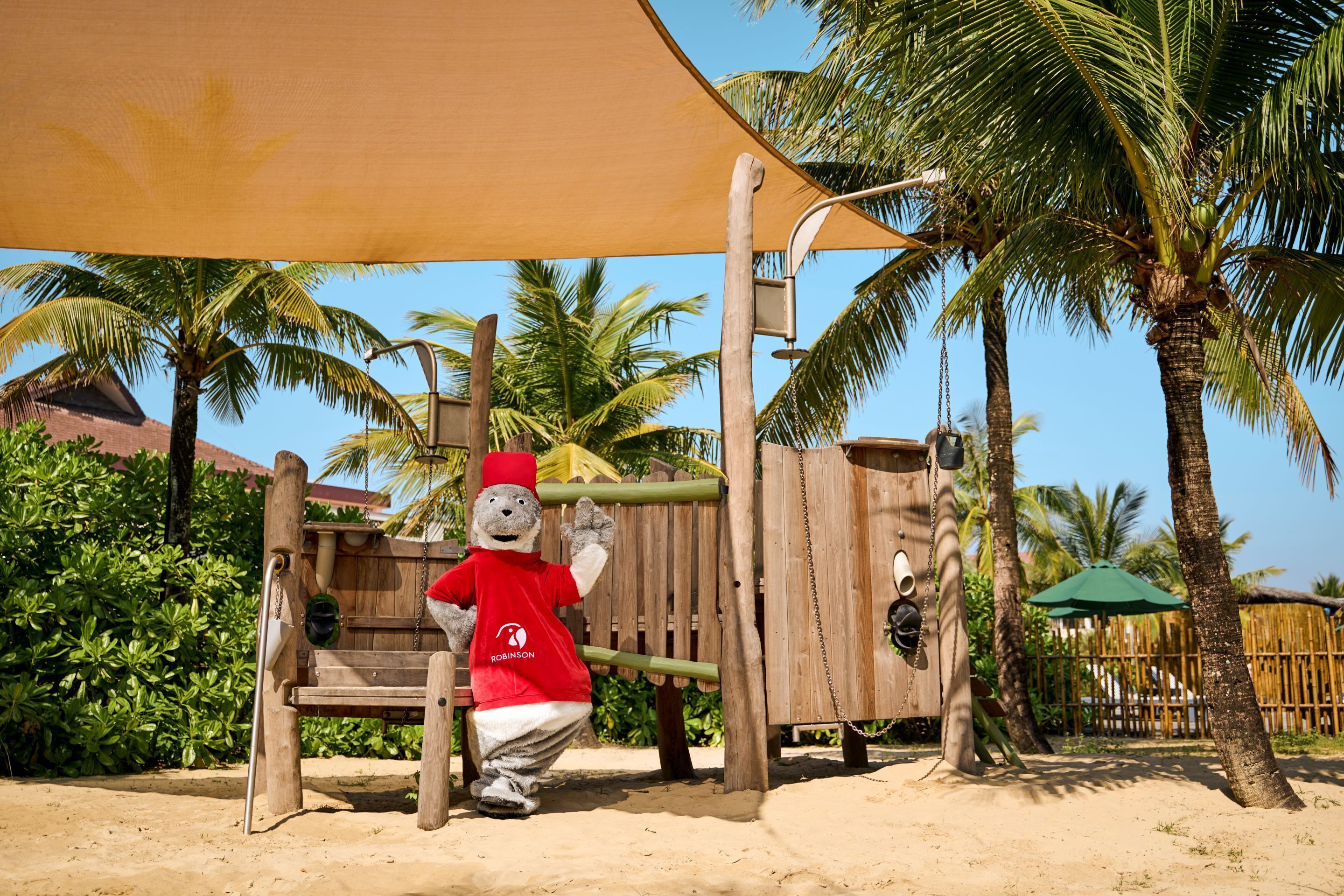 A mascot in a red shirt and hat, a wooden play structure, and palm trees.