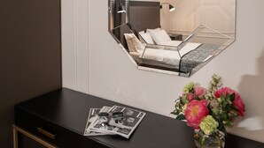 A modern hotel room with a geometric mirror, a bedside table with a magazine and a vase of flowers, and a bed with white linens. - De Paris Apartments (Kyiv)