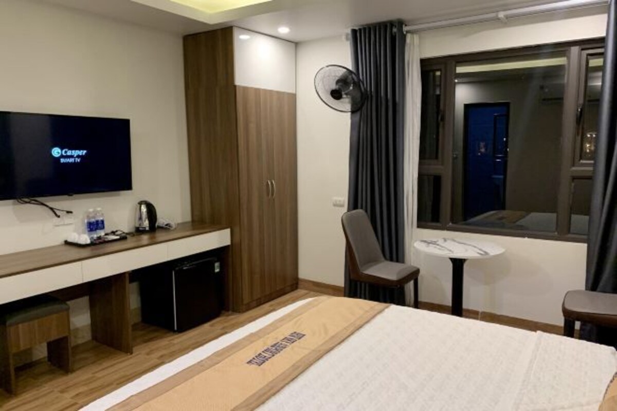 Habitación de hotel con cama, escritorio con televisor, silla, mesita y ventana con cortinas.
