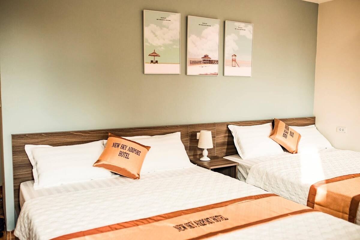 Une chambre d’hôtel avec deux lits, chacun agrémenté d’un oreiller portant l’inscription « NEW SKY AIRPORT HOTEL ». Trois tableaux encadrés sont accrochés au mur au-dessus des lits.