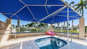 Área da piscina com toldo azul, uma banheira de hidromassagem e vista para palmeiras.