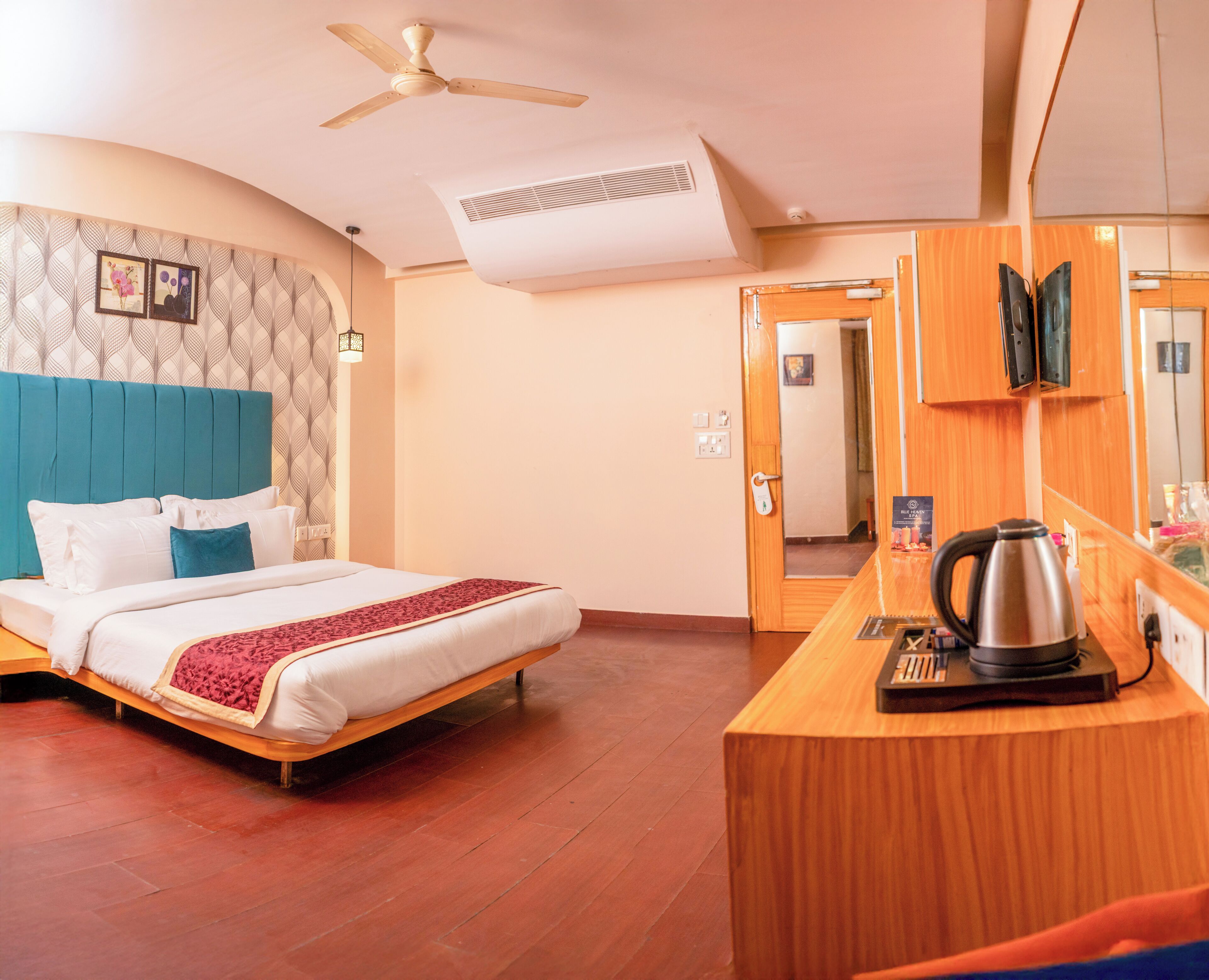 A hotel room with a bed, a desk with a coffee maker, a ceiling fan, and a door leading to another room.