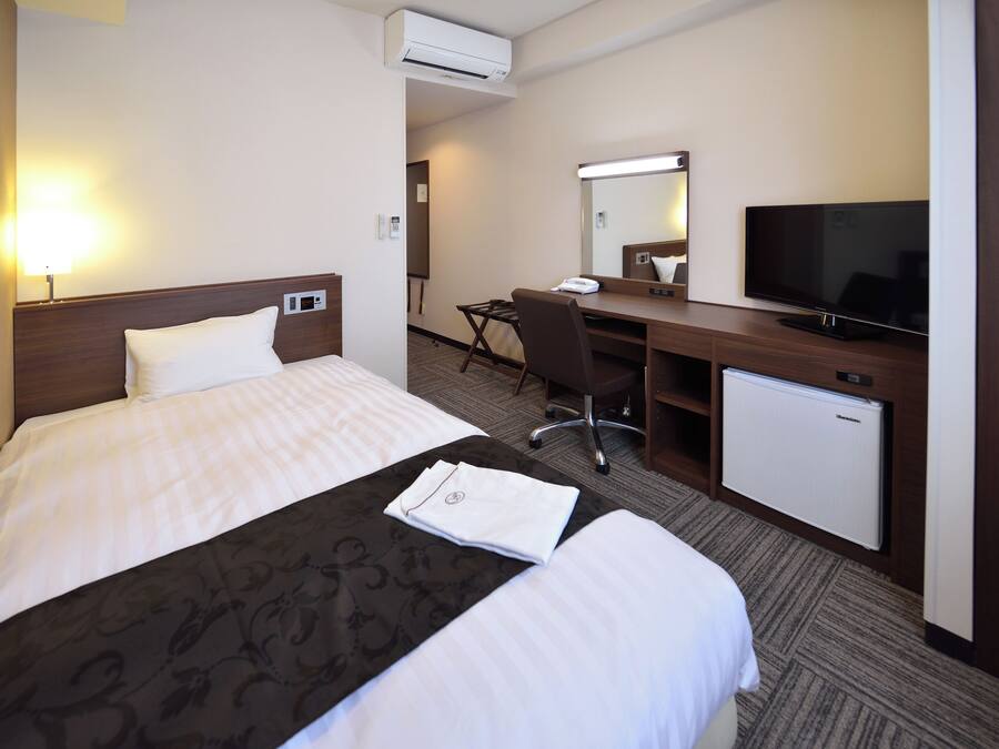 A hotel room with a bed, a desk, a television, and an air conditioning unit.