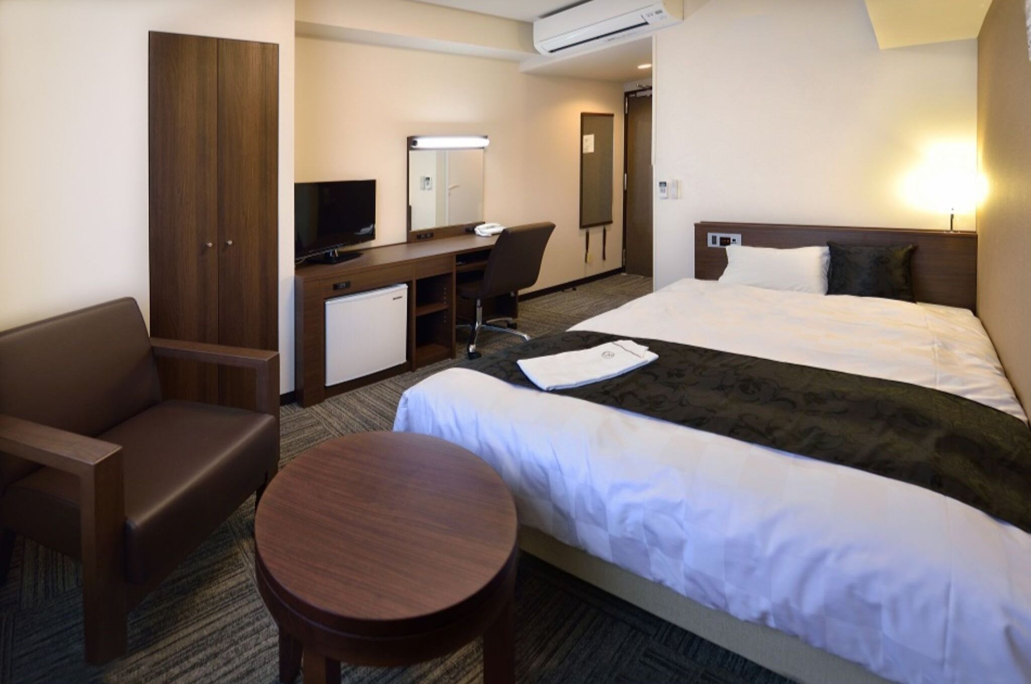 A hotel room with a bed, a desk with a computer, a chair, a small table, and a wardrobe.