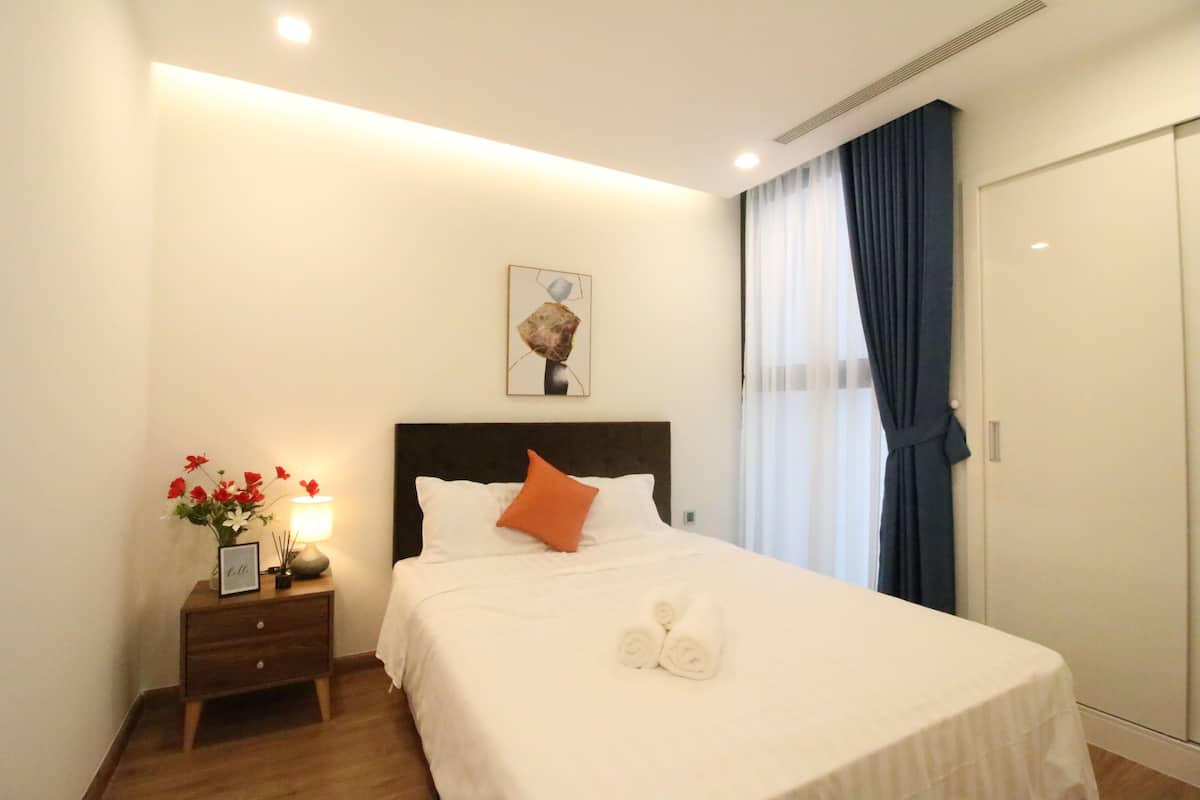 Un lit bien fait, avec du linge de lit blanc et un seul oreiller orange, une table de chevet avec une lampe et un vase de fleurs, et une œuvre d’art encadrée au mur.