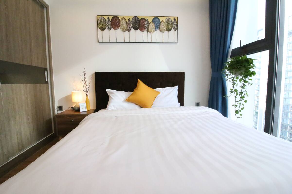 Une chambre d’hôtel comprenant un lit, une table de chevet, une œuvre d’art accrochée au mur, une fenêtre avec des rideaux et une plante.
