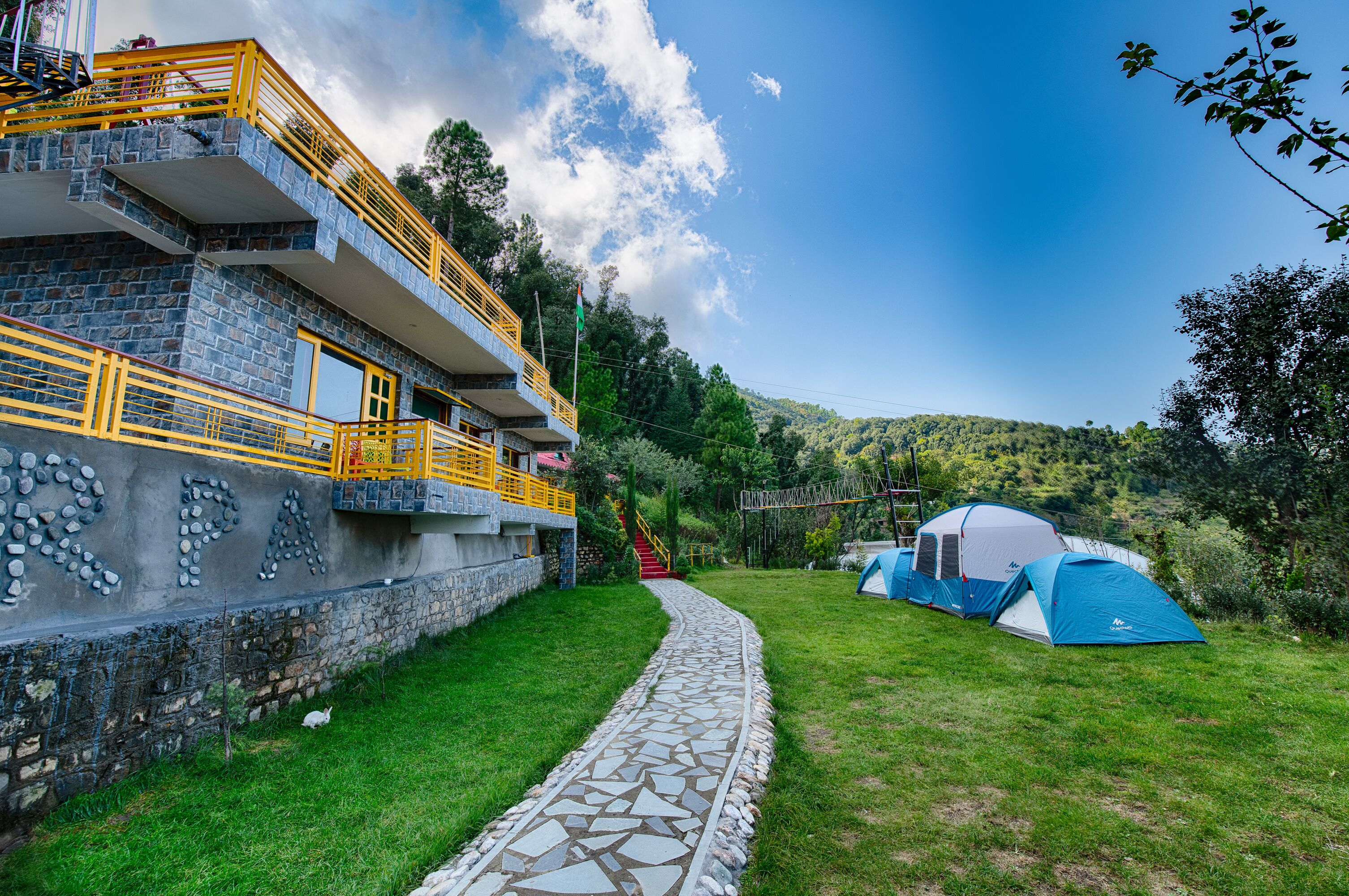Ein Steinhaus mit gelander, ein Steinweg und zwei Zelte auf einer Wiese.