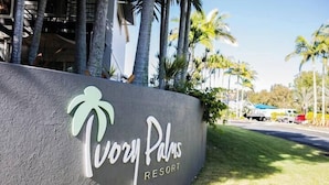 A sign for Ivory Palms Resort with a palm tree logo, surrounded by palm trees and a clear sky. - Unit 88 - Tropical Getaway in 2 Bedroom Unit in 4* Resort (Noosaville)