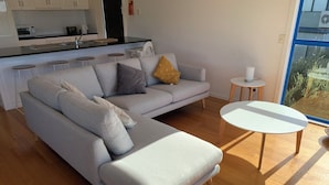 A modern living room with a grey sectional sofa, a small white table, and a view of the kitchen in the background.