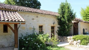 Un bâtiment rustique en pierre, avec un toit de tuiles, des poutres en bois et un petit jardin agrémenté de fleurs blanches.