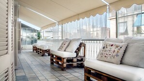 A covered outdoor seating area with wooden benches and cushioned chairs, featuring a decorative railing and large windows.