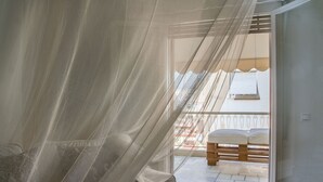 A bedroom with a bed covered by sheer curtains, a window with a view of a balcony with a wooden bench.