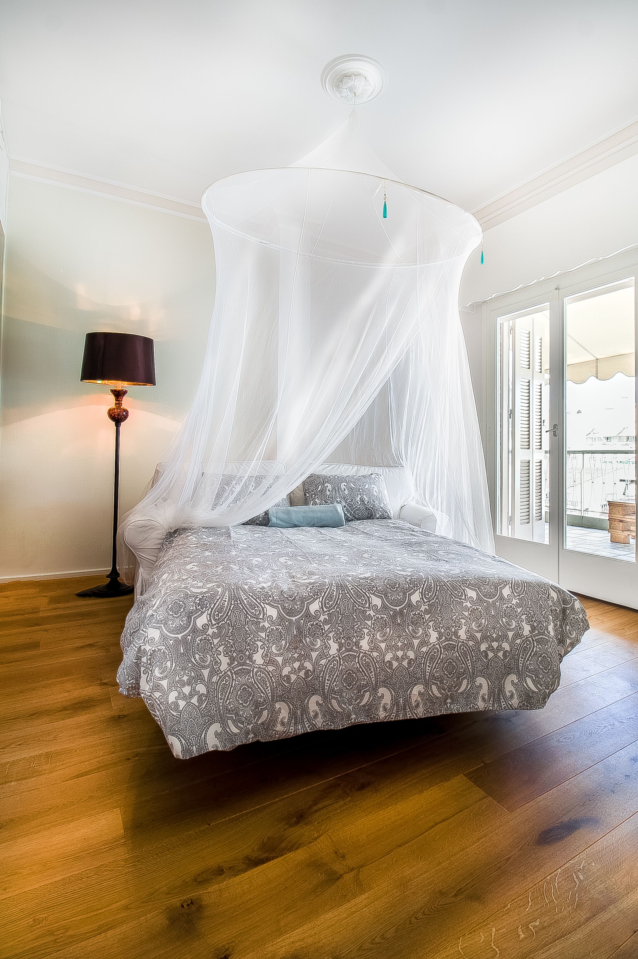 A bedroom with a bed under a canopy, a floor lamp, and a view of the outside through a window.