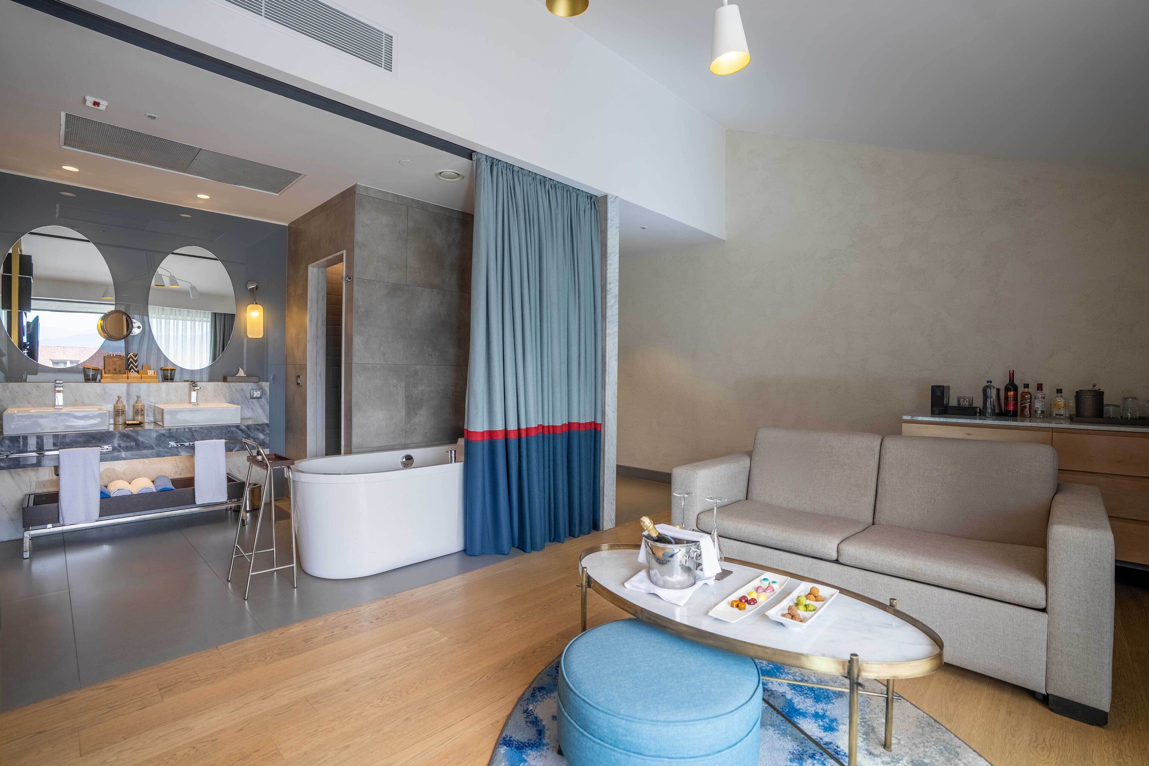A modern hotel room with a bathroom, a sofa, a coffee table, and a dining area.