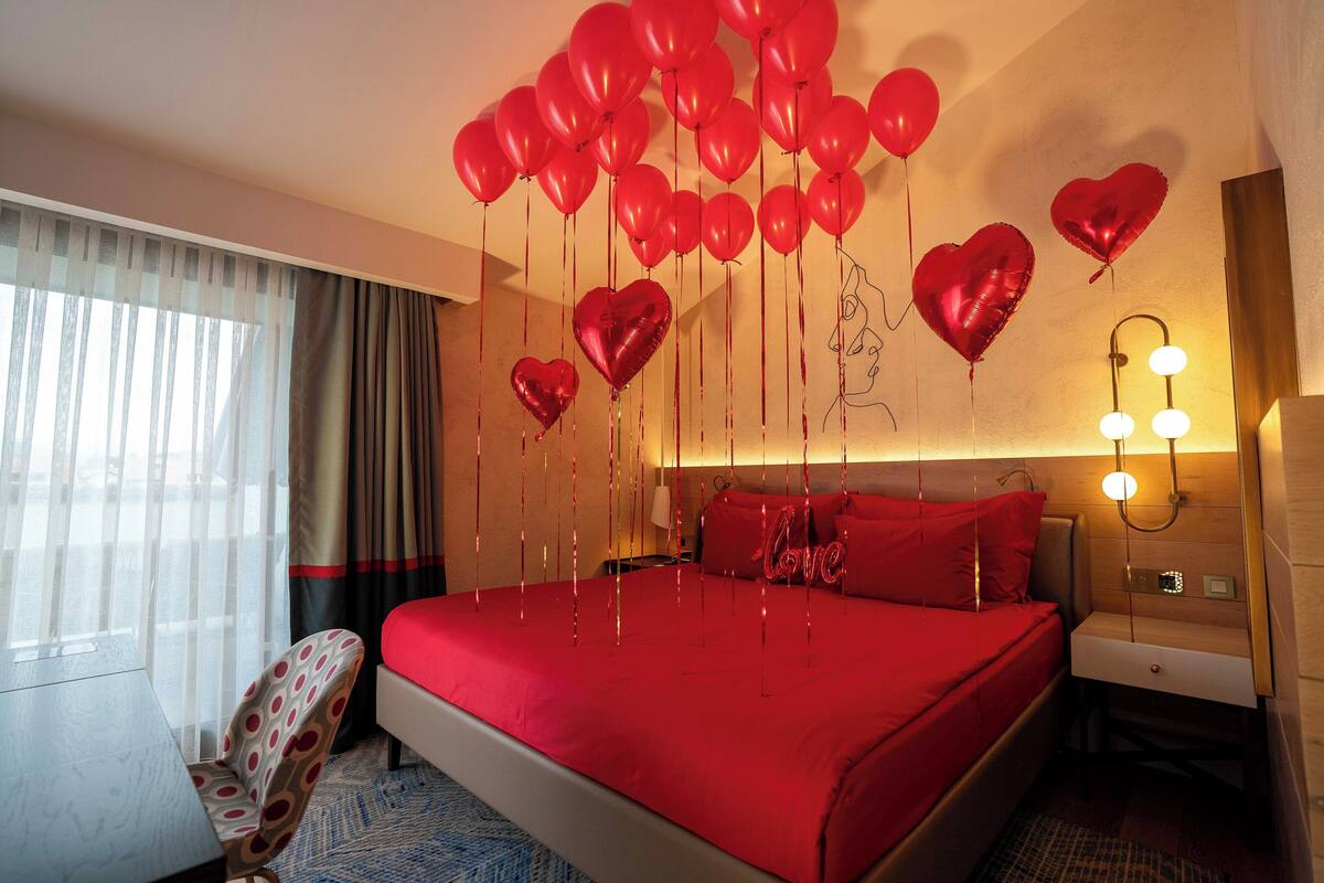 A hotel room with a large bed, red bedding, a chair, and heart-shaped balloons.