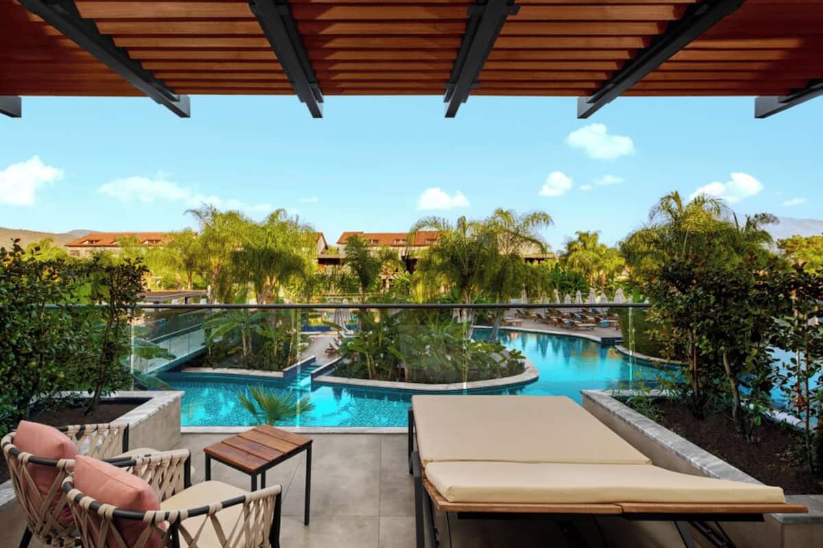 A pool area with lounge chairs and a table, surrounded by lush greenery and a clear blue sky.