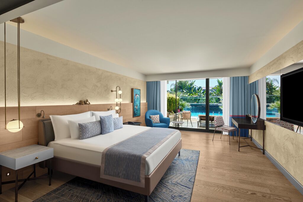 Une chambre d’hôtel moderne, dotée d’un grand lit, d’un bureau, d’une chaise et d’une télévision. La chambre dispose d’une grande fenêtre donnant sur une piscine et un espace verdoyant.