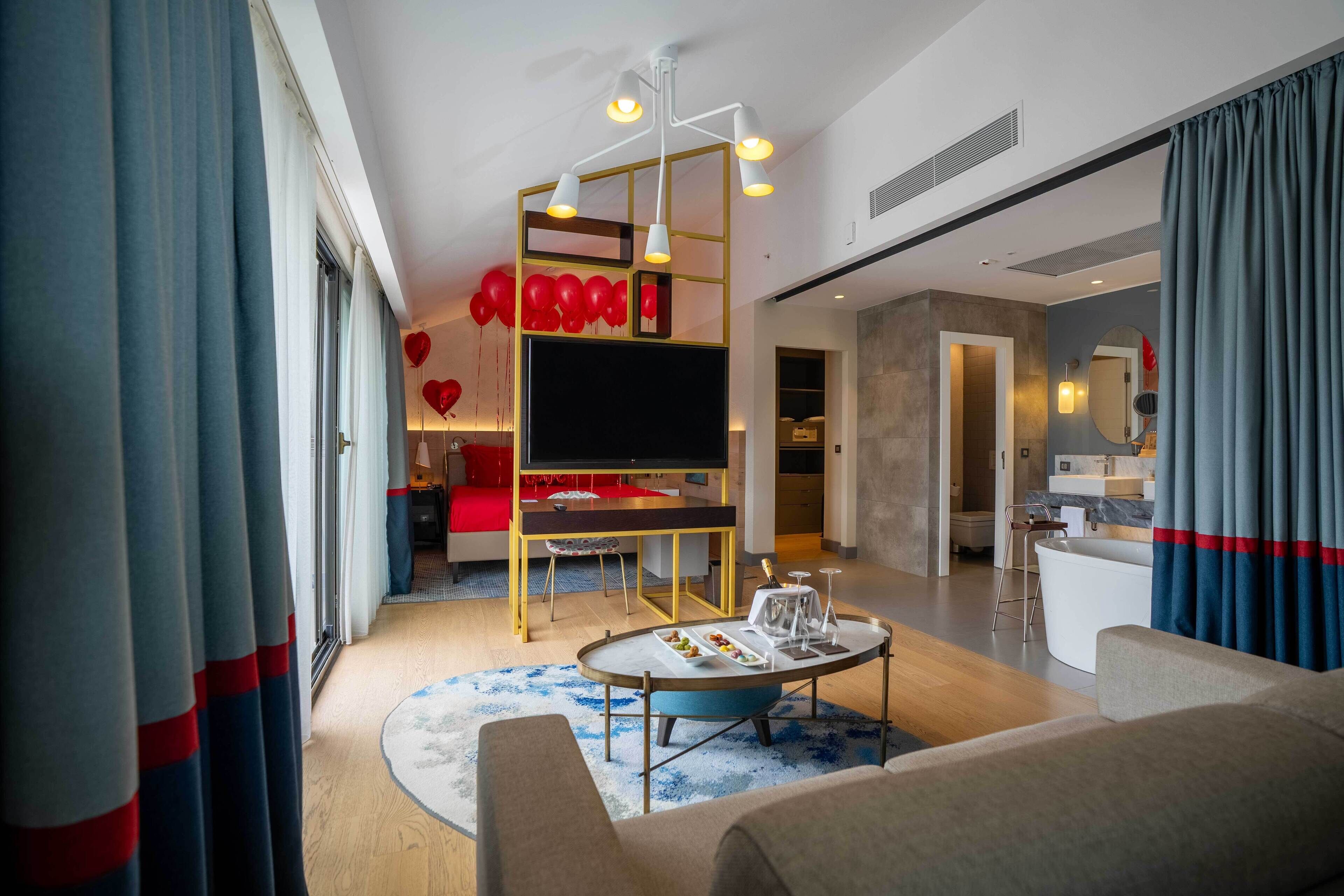 A modern living room with a flat-screen TV, a coffee table, and a sofa. There is a dining area with a red table and chairs. The room has a large window with curtains.