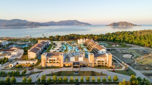 Vista aérea de un resort con varias edificaciones, un área de piscina y una gran extensión de agua con montañas al fondo.
