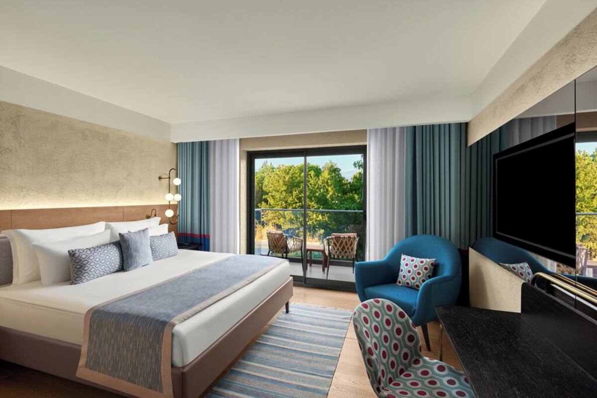 A hotel room with a large bed, a TV, a seating area with a chair, and a balcony with a view of trees.